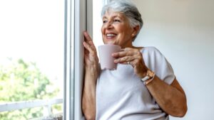 A smiling elderly woman holding a mug looking out the window